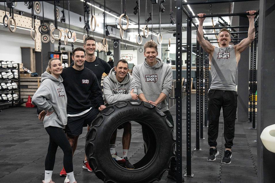 Sechsk&ouml;pfiges Fitness-Team posiert l&auml;chelnd vor einem gro&szlig;en Reifen in einem modernen Trainingsstudio.