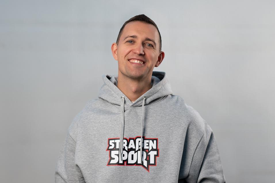 L&auml;chelnder Mann in grauem Hoodie mit dem Schriftzug &bdquo;STRASSEN SPORT&ldquo;, vor einem grauen Hintergrund.