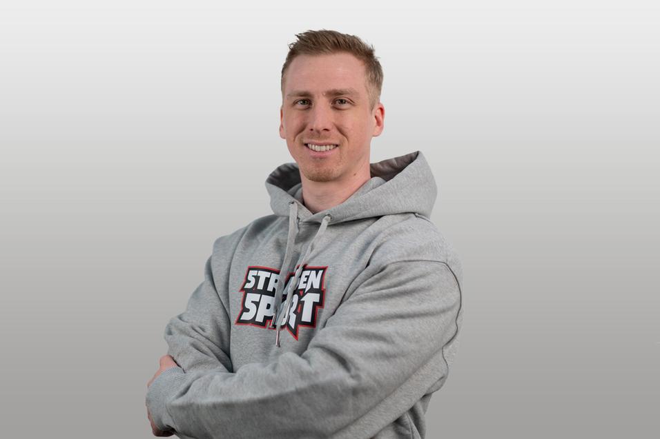 Lächelnder Mann in grauem Hoodie mit "STRENGEN SPORT"-Aufdruck, verschränkt die Arme vor einem neutralen Hintergrund.