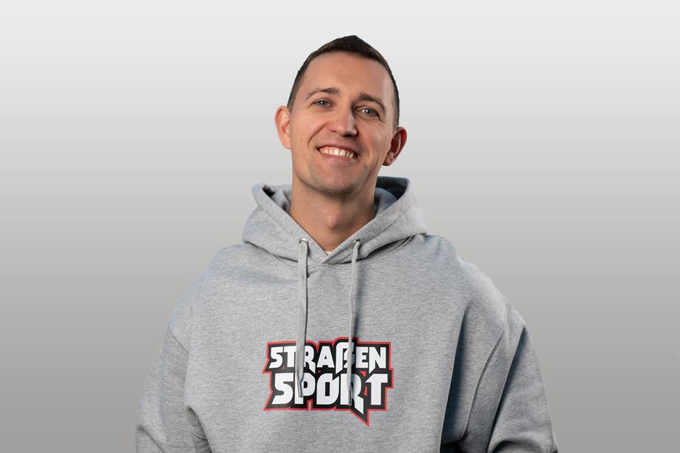 Junger Mann mit kurzem Haar und grauem Hoodie, lächelt vor neutralem Hintergrund. Auf dem Hoodie steht „STRASSEN SPORT“.