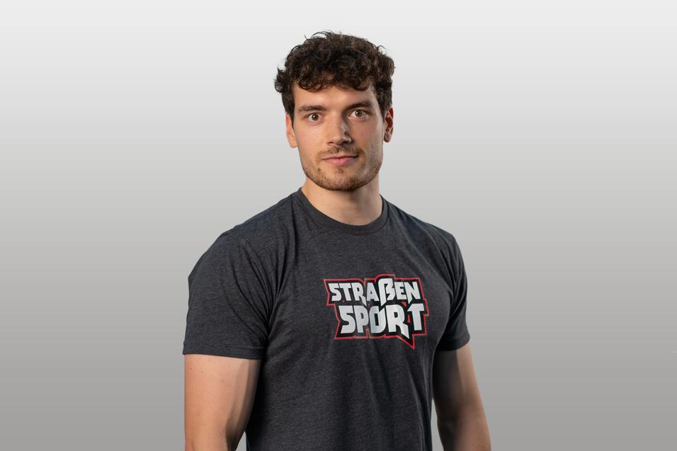 Junger Mann mit lockigem Haar in grauem T-Shirt mit dem Schriftzug "STRAßENSPORT" vor neutralem Hintergrund.
