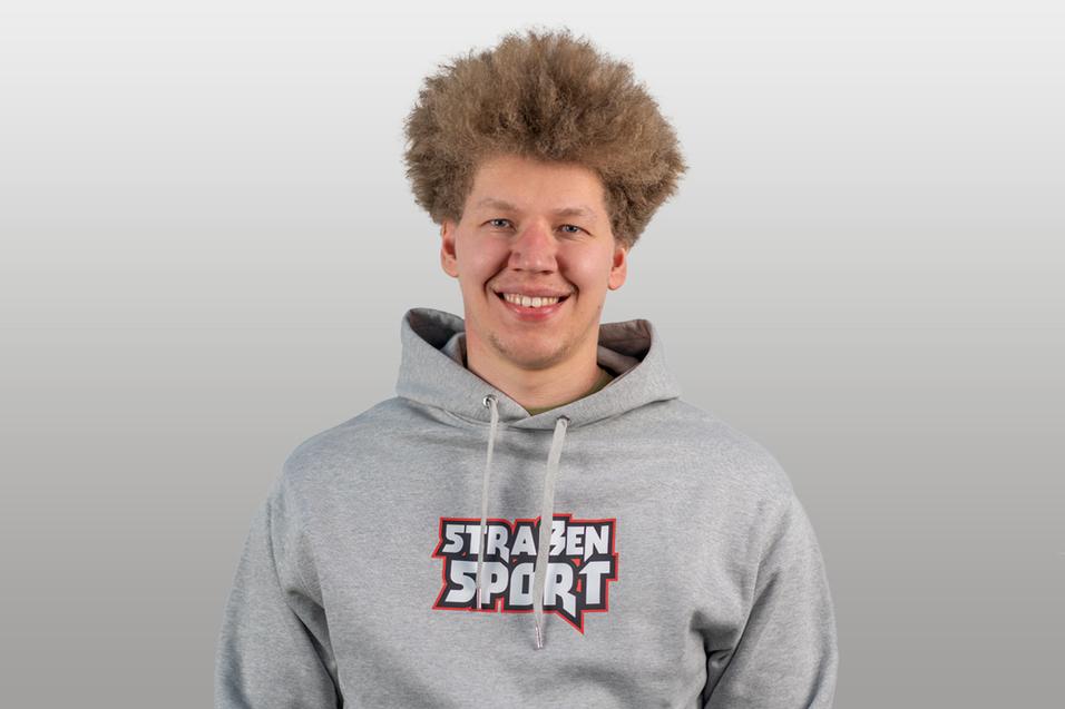 Junger Mann mit lockigem Haar trägt einen grauen Hoodie mit dem Aufdruck „STRAßENSPORT“. Er lächelt in die Kamera.