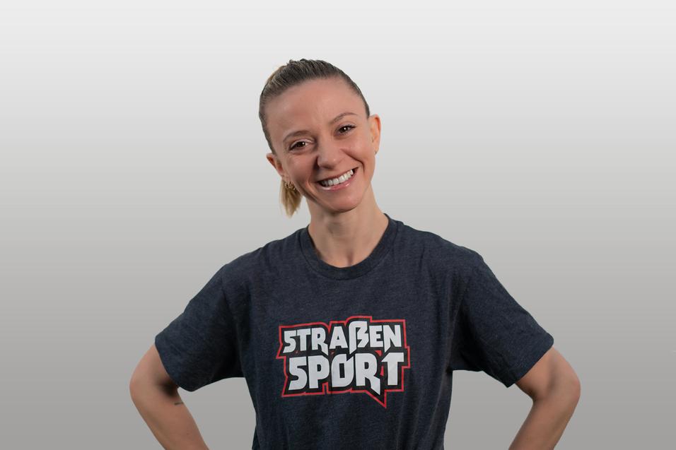 Lächelnde Person mit kurzem Haar in grauem T-Shirt mit dem Aufdruck "STRAßEN SPORT". Hintergrund in sanften Grautönen.