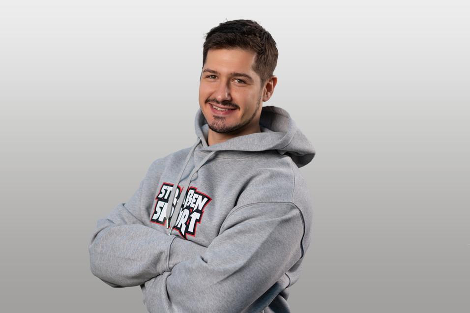 Junger Mann mit lässigem Lächeln, trägt grauen Hoodie und steht vor einem neutralen Hintergrund.