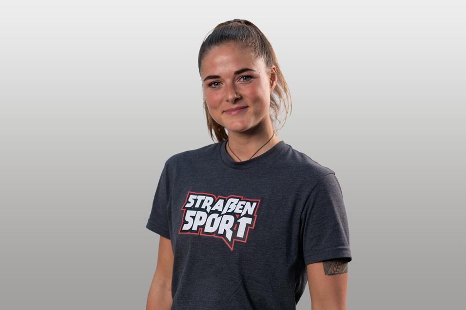 Porträt einer jungen Frau in einem grauen T-Shirt mit dem Aufdruck „STRAßENSPORT“, mit lächelndem Gesicht.