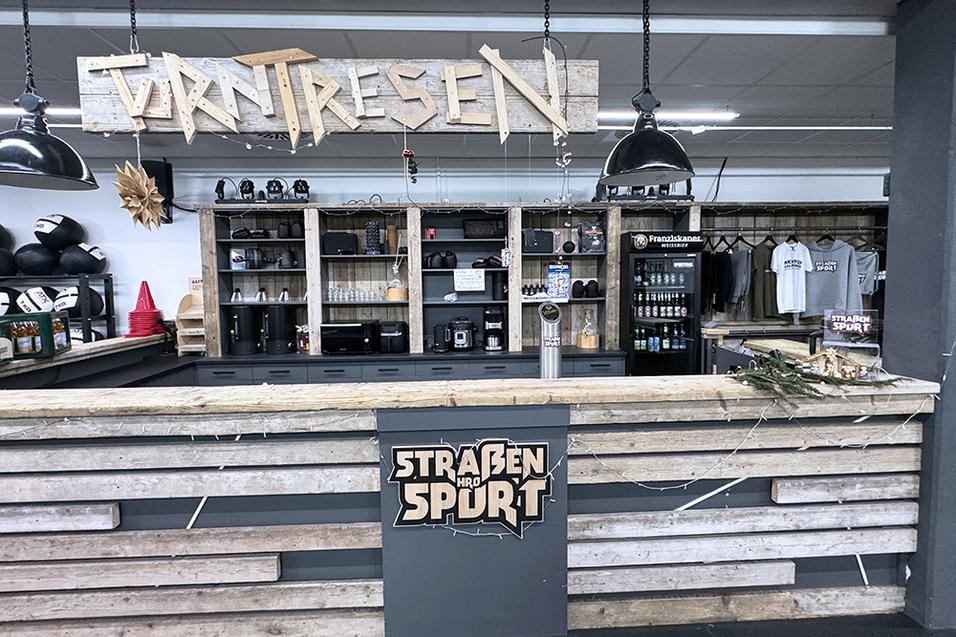 Theke in Sporteinrichtung mit Holzverkleidung, Hängelampen und Merchandise-Artikeln im Hintergrund.