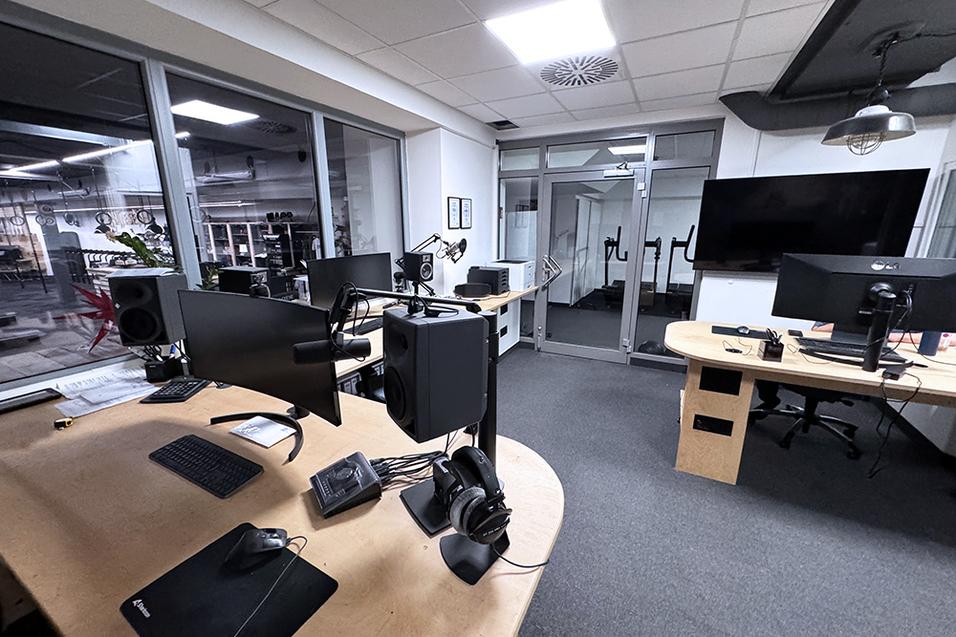 Büroansicht mit zwei Monitoren, Lautsprechern und Headset auf einem Tisch; im Hintergrund ein großer Fernseher.