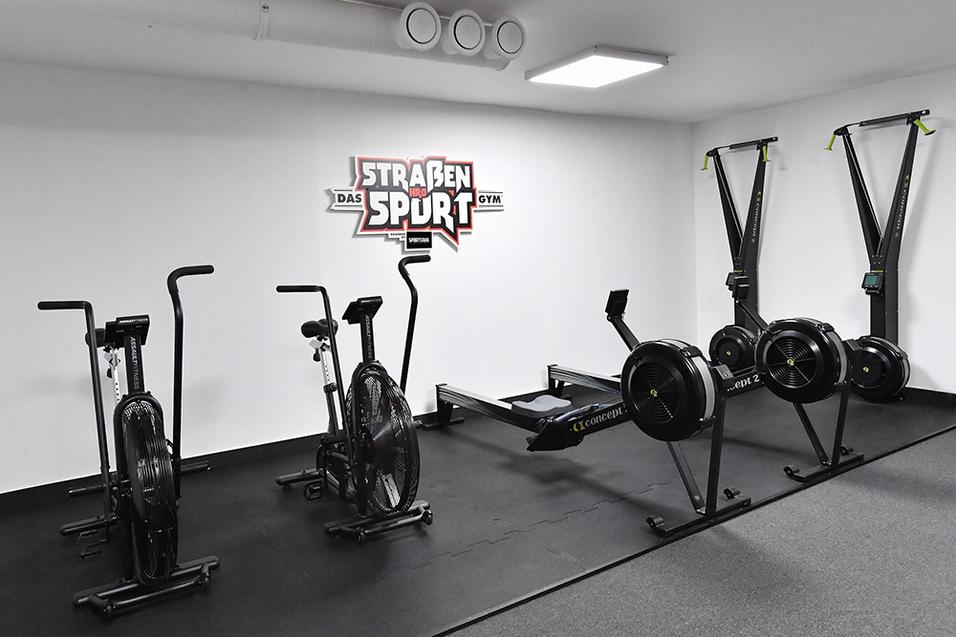 Fitnessraum mit verschiedenen Ergometern, Rudergeräten und einem Wandlogo von "Straßen Sport Gym".