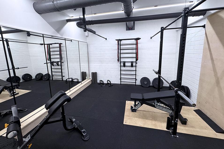 Fitnessraum mit Geräten, Spiegel und Gewichten auf schwarzem Bodenbelag.
