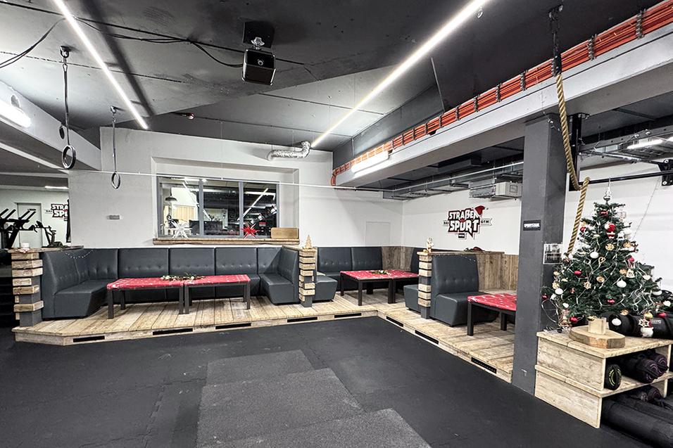 Gemütlicher Sitzbereich mit einer L-förmigen Couch, Tischen und einem Weihnachtsbaum in einem Fitnessstudio.