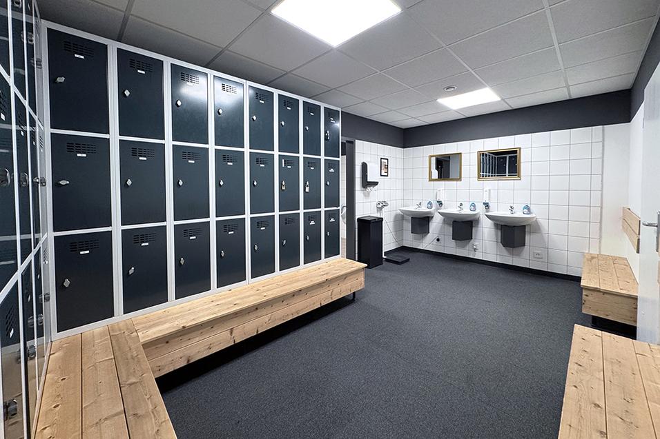 Umkleideraum mit schwarzen Spinden, Holzbank, Waschbecken und Spiegeln an den Wänden. Warmer, neutraler Farbton.