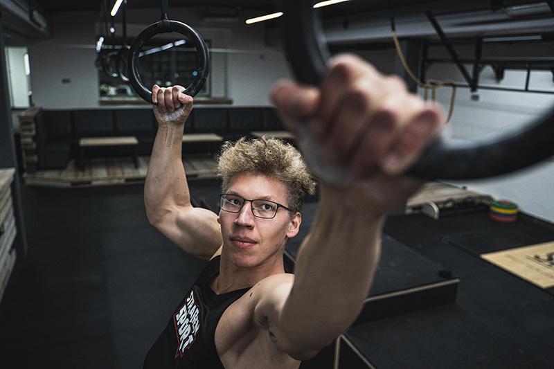 Junger Mann mit Brille, der an Turnringen hängt, im Hintergrund eine moderne Trainingsumgebung.