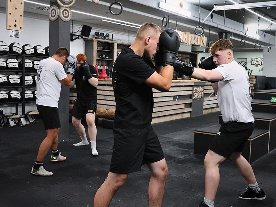 Vier junge Männer trainieren Boxen in einem Fitnessstudio, angelehnt an verschiedene Trainingsgeräte und Boxsäcke.
