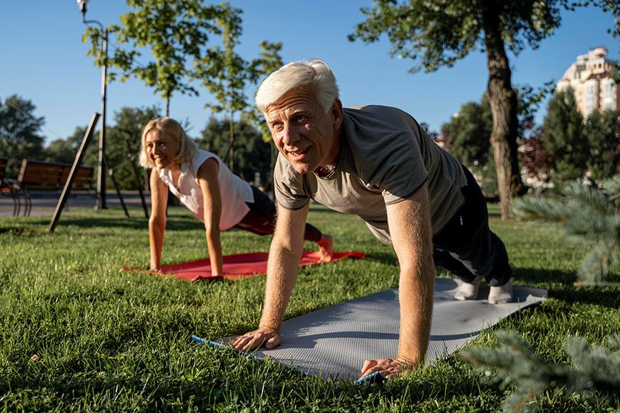 Zwei ältere Personen machen im Park Liegestütze auf Yogamatten im Grünen, umgeben von Bäumen und einem sonnigen Ambiente.
