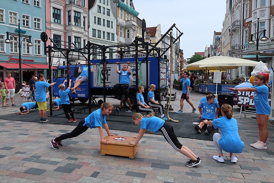 Gruppe von Kindern in blauen T-Shirts beim Sporttraining auf einer Straßenfläche mit Fitnessgeräten und Zuschauern.