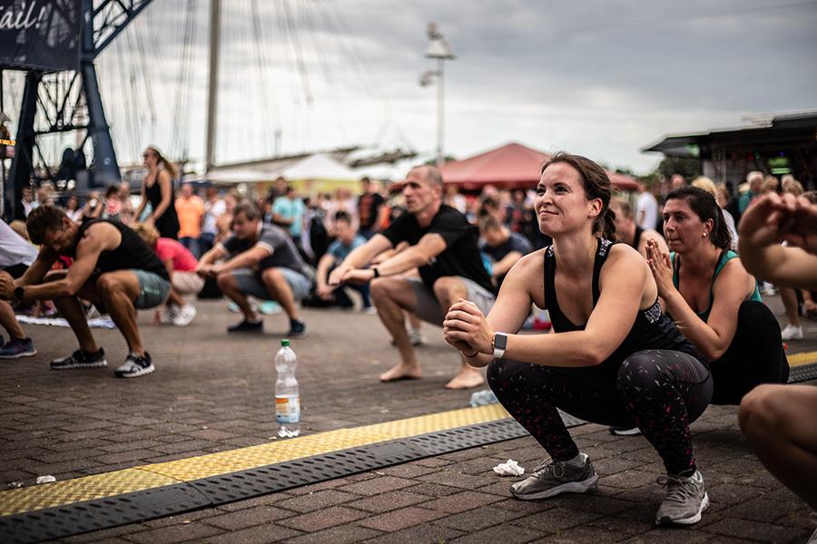 Gruppe von Sportlern macht Squats im Freien, mit Zuschauern und einem überdachten Bereich im Hintergrund.