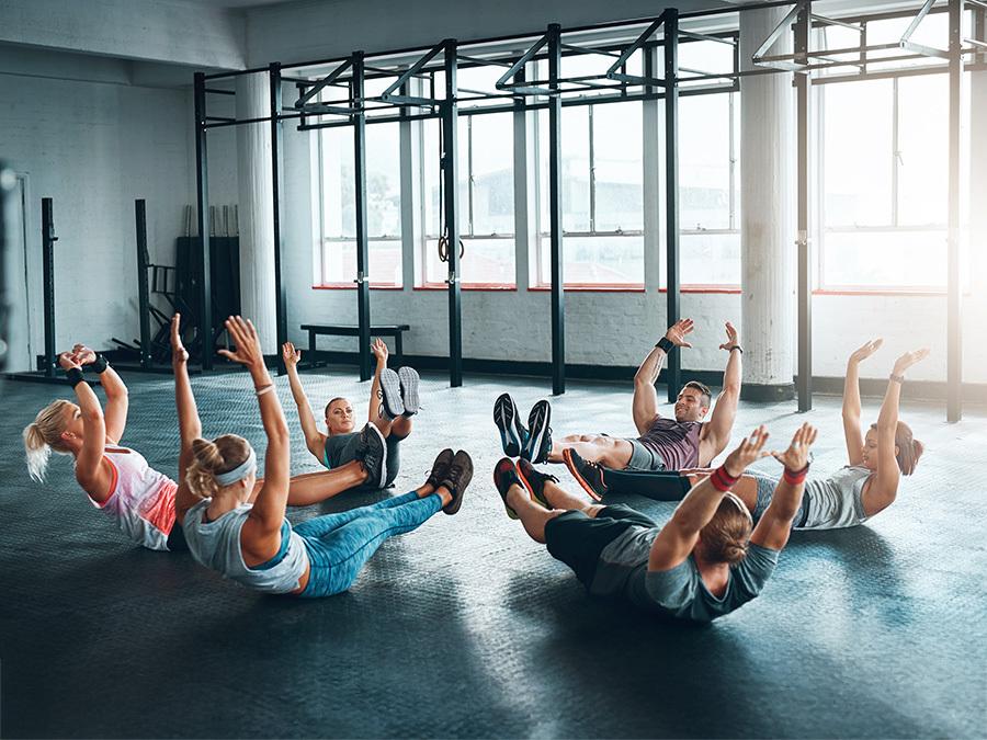 Gruppe von sechs Personen, die in einem Fitnessstudio sitzende Rumpfbeugen ausf&uuml;hren. Helle Fenster im Hintergrund.