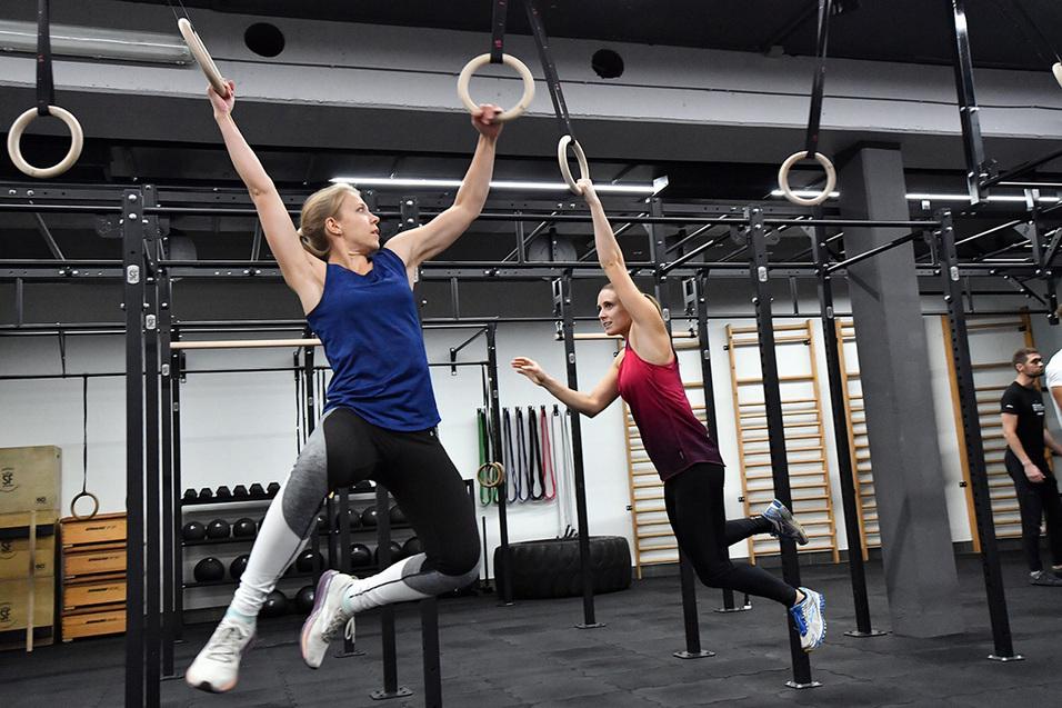 Zwei Frauen trainieren an Gymnastikringen in einem Fitnessstudio, eine von ihnen springt und greift nach einem Ring.