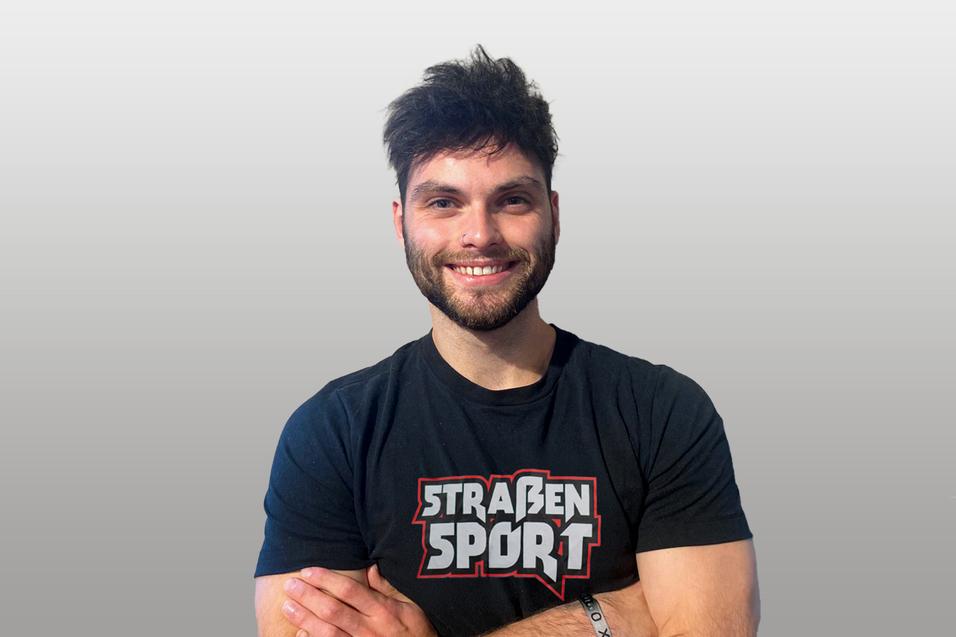 Mann mit kurzen Haaren und Bart lächelt, trägt ein schwarzes T-Shirt mit dem Aufdruck „STRASSENSPORT“.