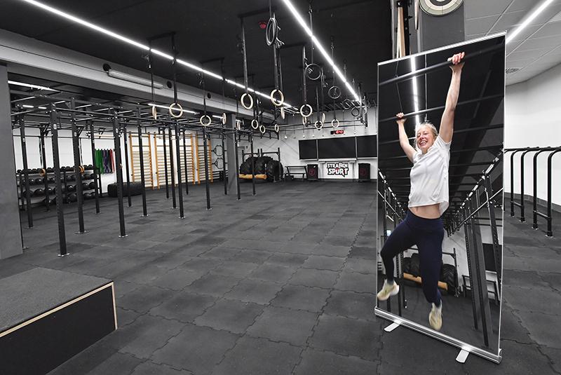 Frau hängt an Klimmzugstange vor einer modernen Fitnessstudio-Ausstattung mit Trainingsgeräten im Hintergrund.
