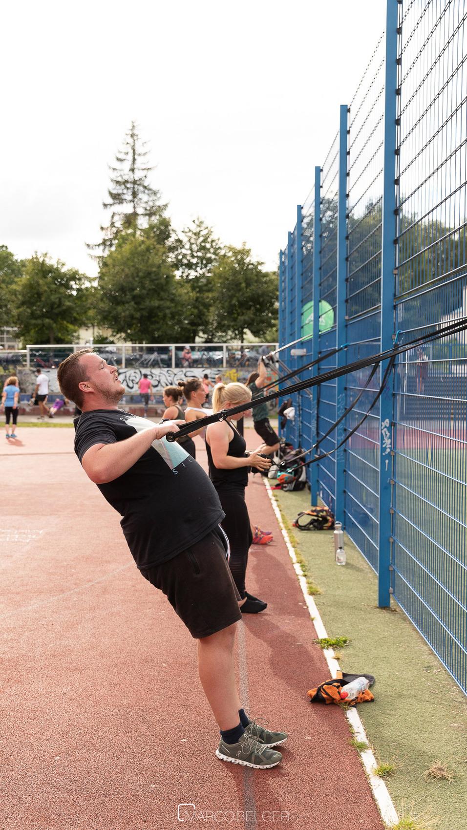 Mann trainiert mit Widerstandsband an einer blauen Wand, w&auml;hrend andere Sportlerinnen im Hintergrund &Uuml;bungen machen.