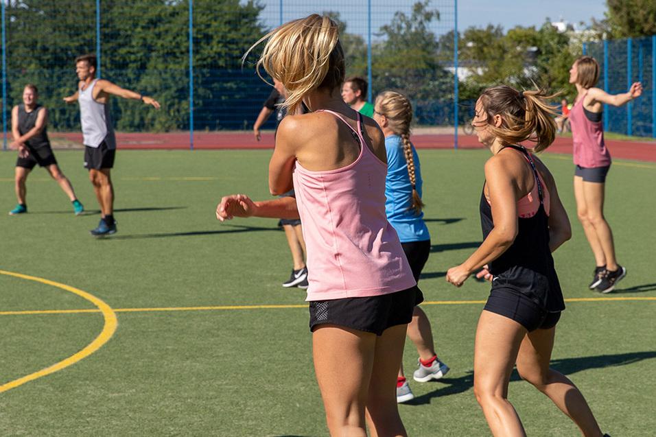 Gruppe von sechs Personen in Sportkleidung beim Training auf einem Kunstrasenplatz an einem sonnigen Tag.