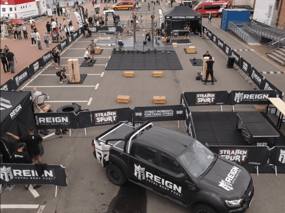 Blick auf ein Fitness-Event im Freien mit mehreren Aktivit&auml;ten, Teilnehmern und Sponsorenst&auml;nden auf einem Parkplatz.